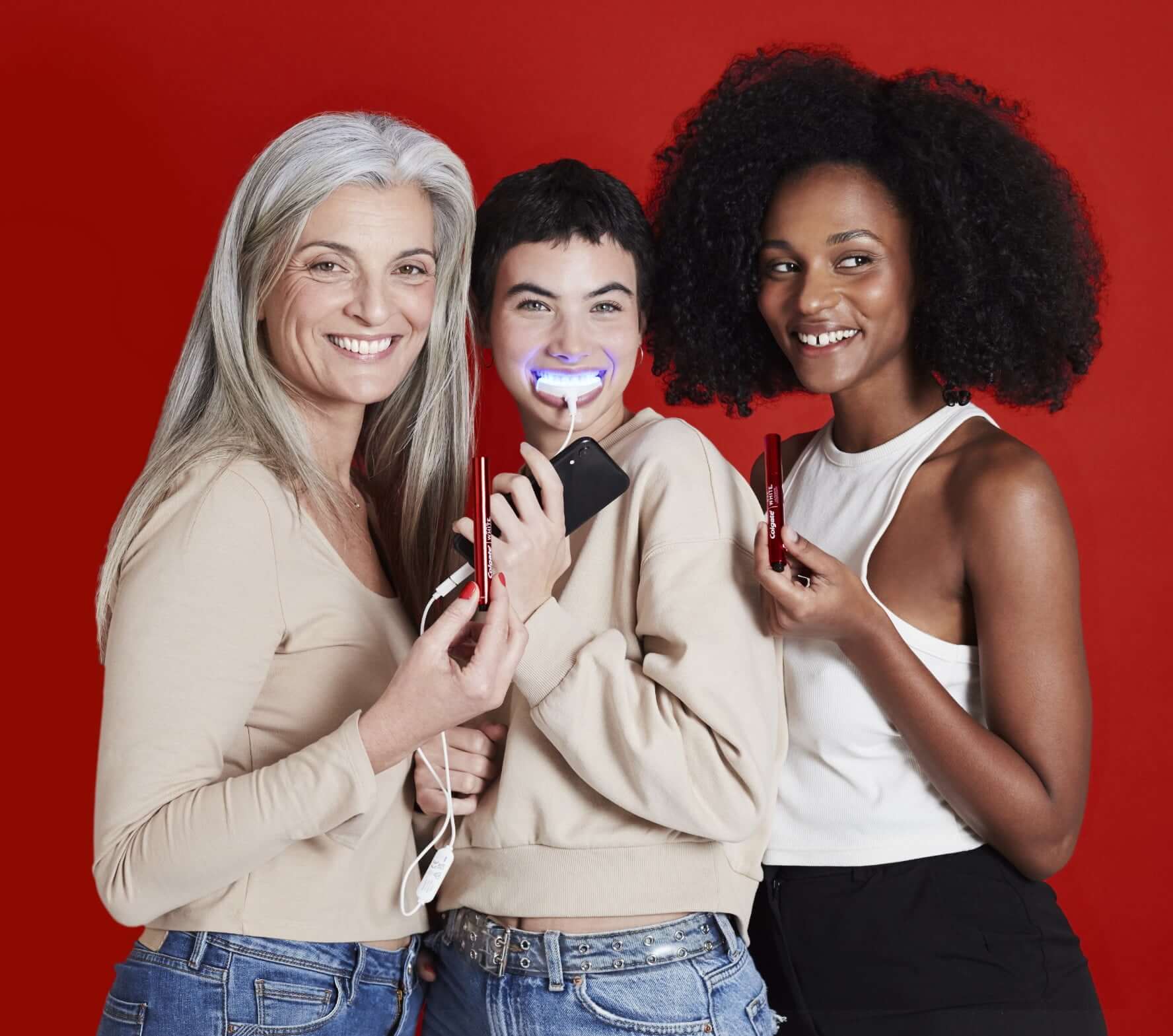 Drei Frauen vor einem roten Hintergrund halten den Colgate Whitening Stift und das Whitening Kit in der Hand